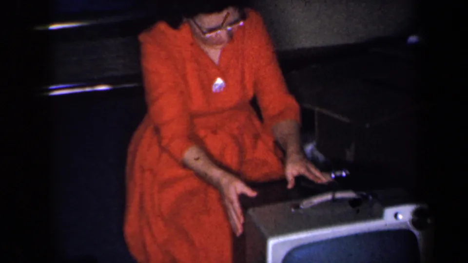 In 1959 New York home-movie footage, a woman in glasses and a red dress reaches toward controls on a vintage television set.