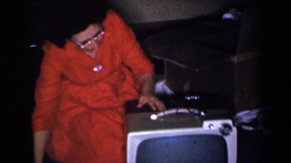 The woman in a red dress and glasses bends over the television cabinet, with one hand near the top controls in the private room.