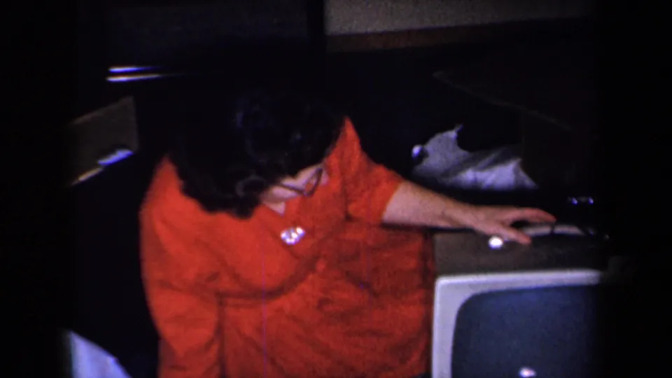 A high home-movie angle shows the woman reaching across the top of the vintage television, with the screen and nearby furniture visible.