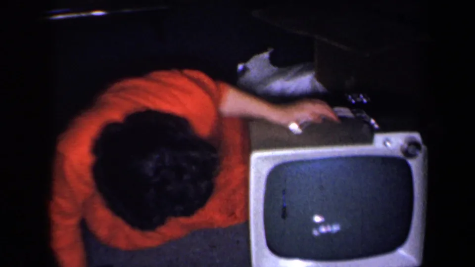 The woman bends low over the television cabinet, her hand near a control knob, as the rounded screen catches a small reflection.