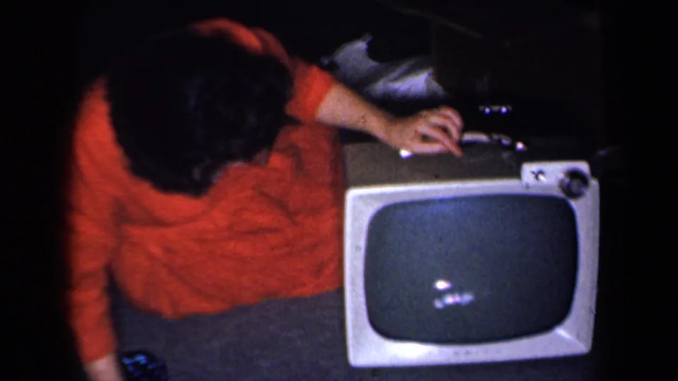 In a dim 1959 New York home interior, a woman in a red dress bends toward a portable television with controls visible.