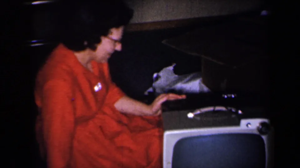 The woman sits beside a portable television in a dim private room, with eyeglasses and red clothing visible.
