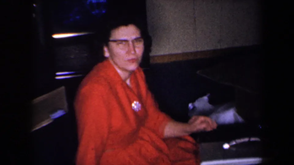 A woman in glasses and a red dress looks toward the camera while seated near a portable television in a New York home-movie frame.