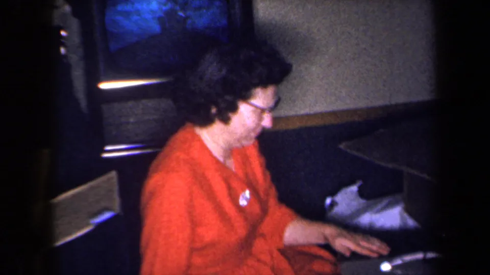 The woman remains seated on the floor near the portable television, her hand resting close to the set in dim home-movie light.