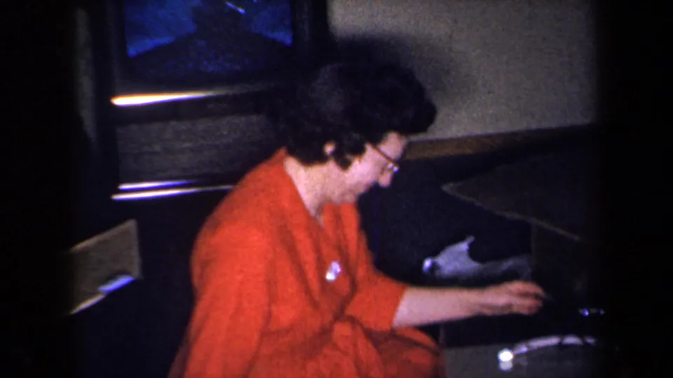 In a 1959 New York home-movie frame, a woman in glasses and a red dress sits near a television in a dim private room.