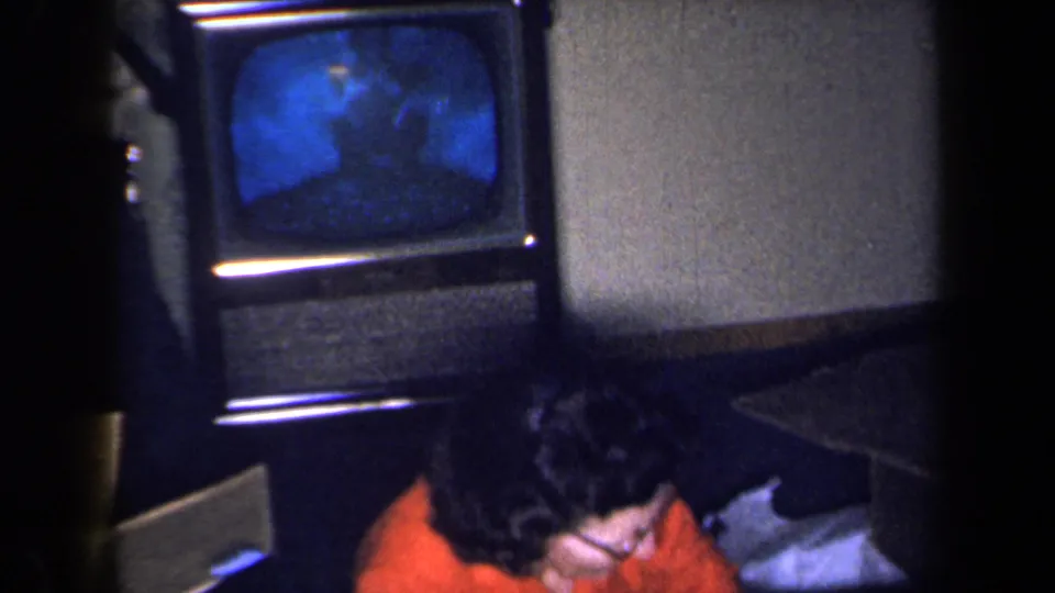 The television screen glows blue in a dim 1959 New York interior as the woman in red leans low near the set.