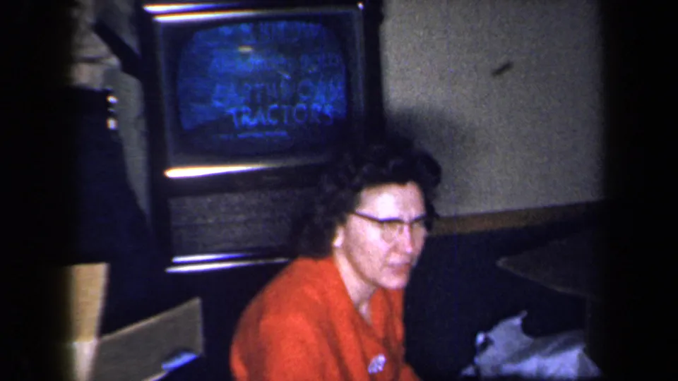 A woman in glasses and a red dress turns toward the camera beside a television in the 1959 New York home-movie setting.