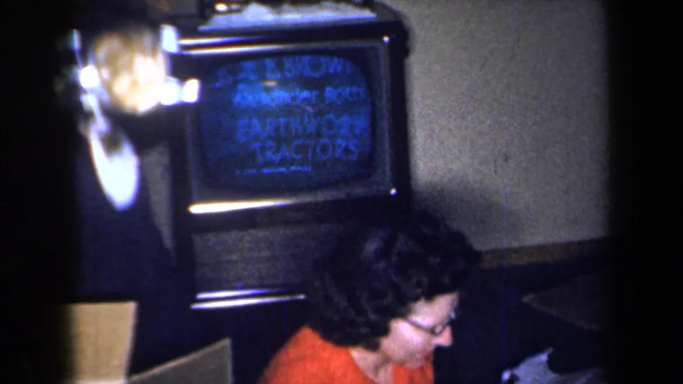 The television shows credits for Earthworm Tractors while the woman in red sits below the screen in a private 1959 New York interior.