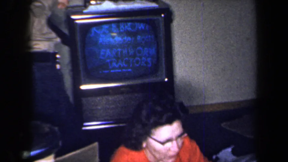 1959 New York home-movie frame showing a woman seated beside a small television with blue text in a dim private interior.