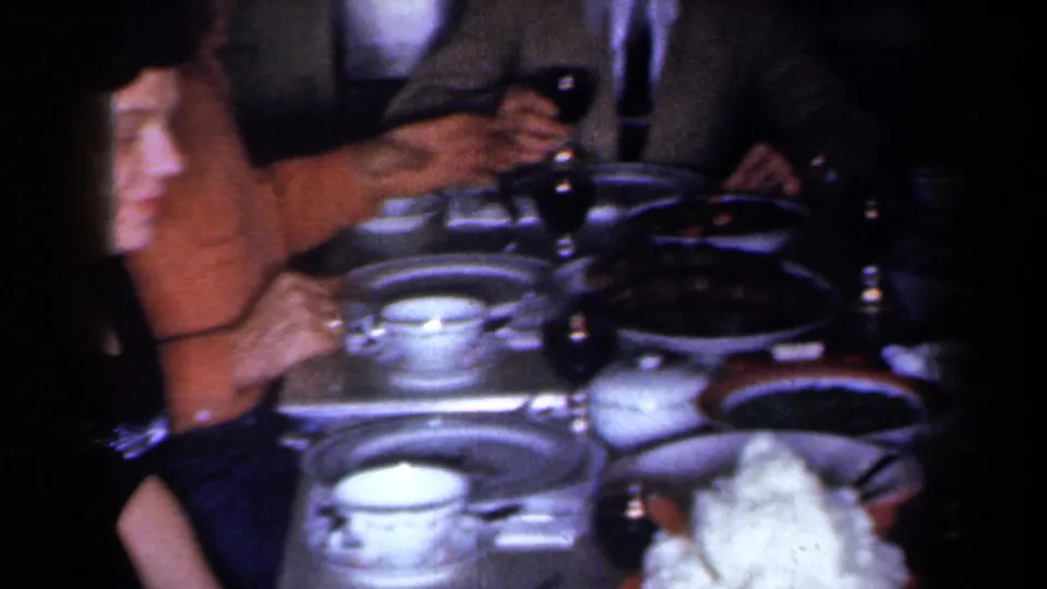 1959 New York home-movie frame showing a woman in orange at a crowded dinner table with china and serving dishes.