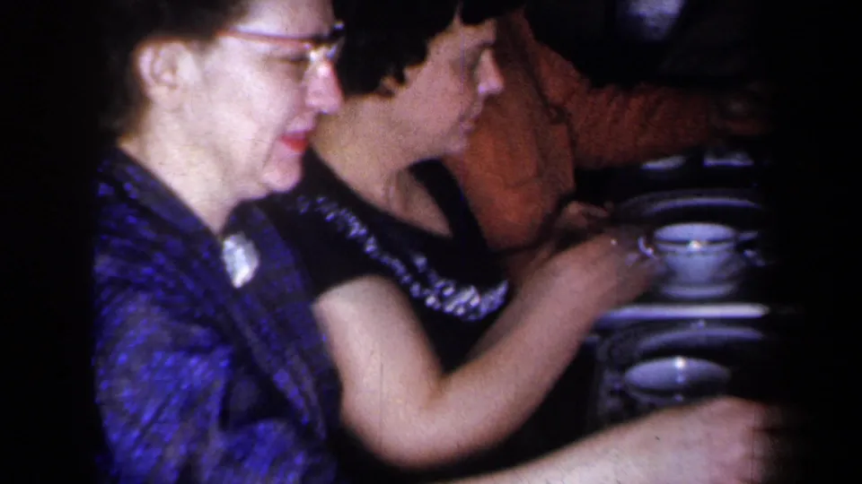 1959 New York home-movie frame of two women seated side by side at a dining table in a dim private interior.