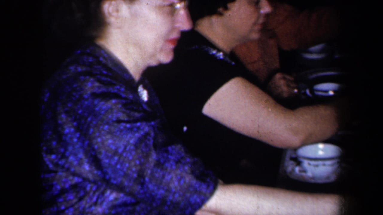 In 1959 New York home-movie footage, two adult women sit close together at a dimly lit dinner table with china and plates nearby.