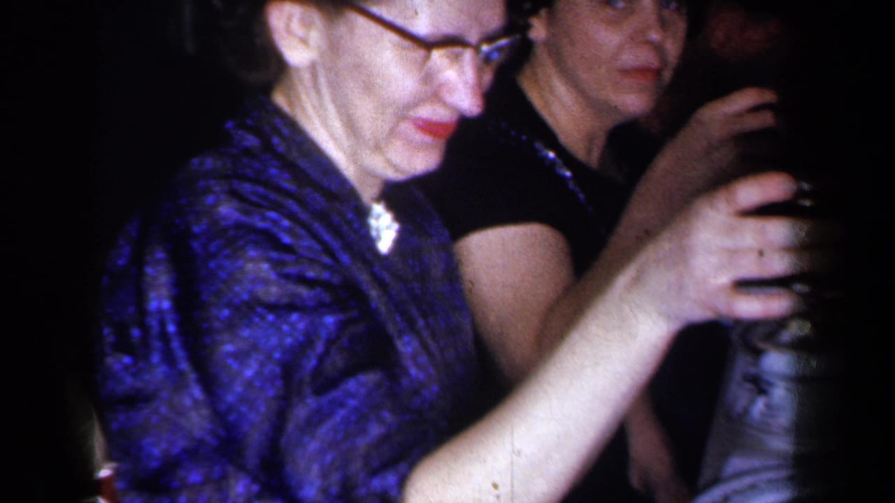 A woman in glasses and a blue patterned dress raises a dark glass at a 1959 New York dinner table, with another adult guest beside her.