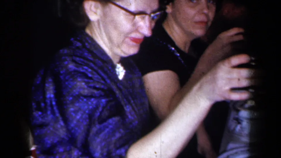 A woman in glasses and a blue patterned dress raises a dark glass at a 1959 New York dinner table, with another adult guest beside her.