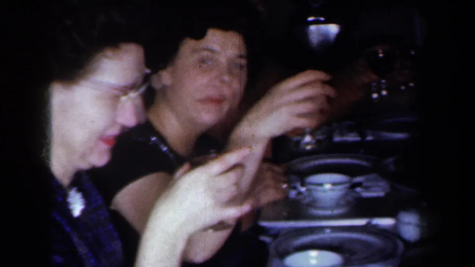 Two adult women sit at a 1959 New York dinner table, one holding a small glass as cups, plates, and tableware fill the foreground.