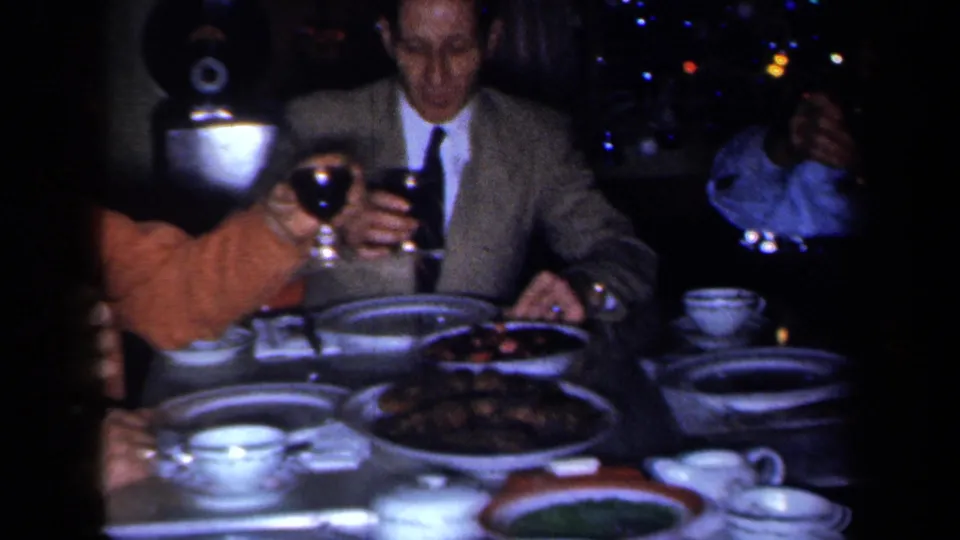 Adult guests in dressy clothing sit around a 1959 New York dinner table with raised wine glasses, china cups, plates, and serving dishes.