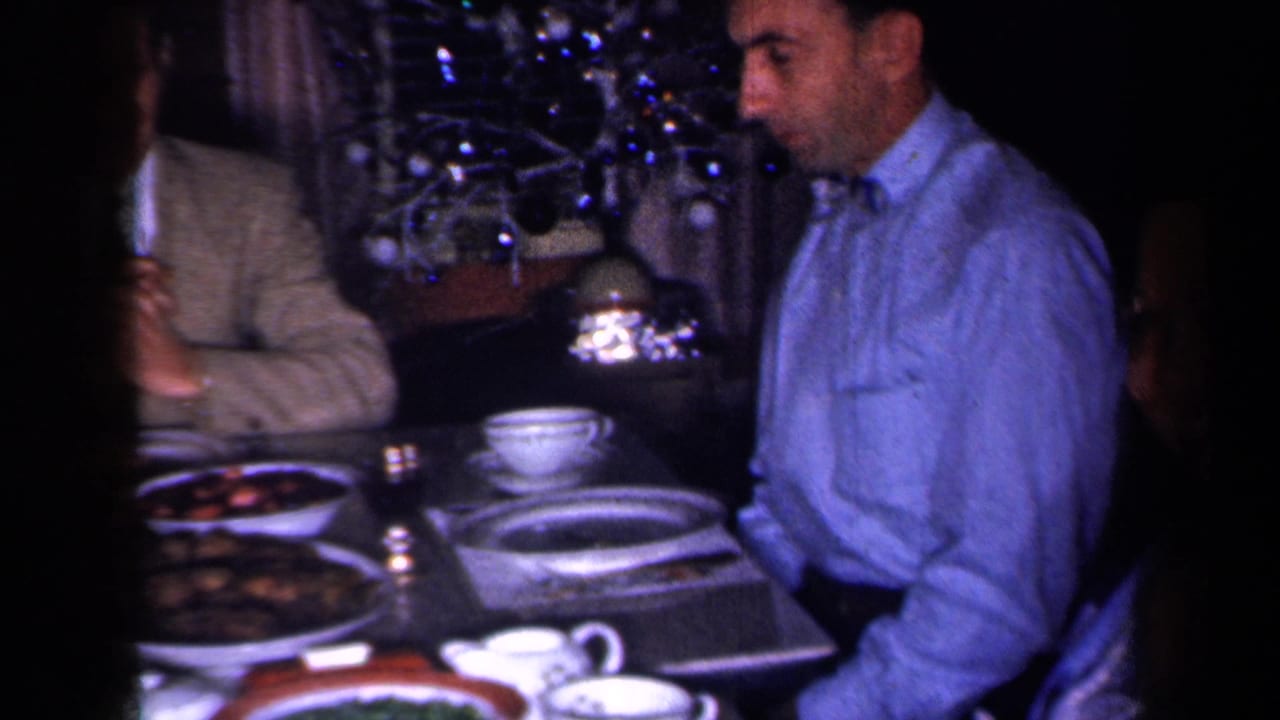Man in a blue shirt sits beside a table with food and cups, with a lit tree behind him.