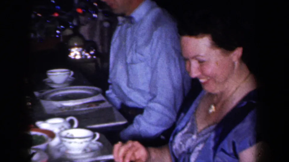 1959 New York home-movie frame showing a smiling woman in formal dress seated at a Christmas dinner table.