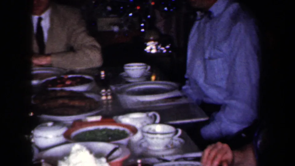 1959 New York Christmas dinner frame with a crowded table, china cups, serving bowls, and adults seated near tree lights.