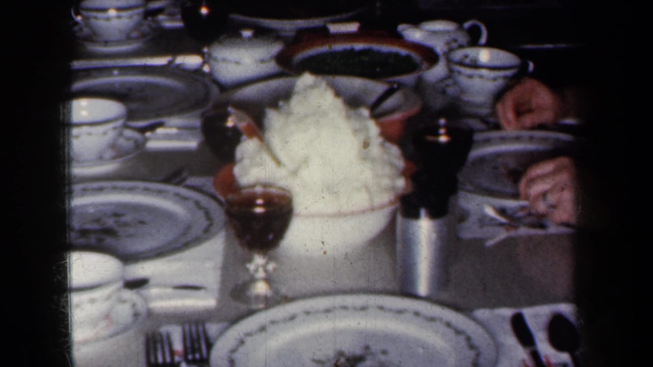 1959 New York home-movie frame showing a formal table spread with china, cups, silverware, stemware, food, and partially visible guests.
