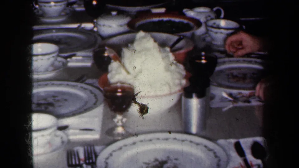 1959 New York home-movie frame showing a formal dinner table with patterned china, cups, wine glasses, and a central serving of potatoes.
