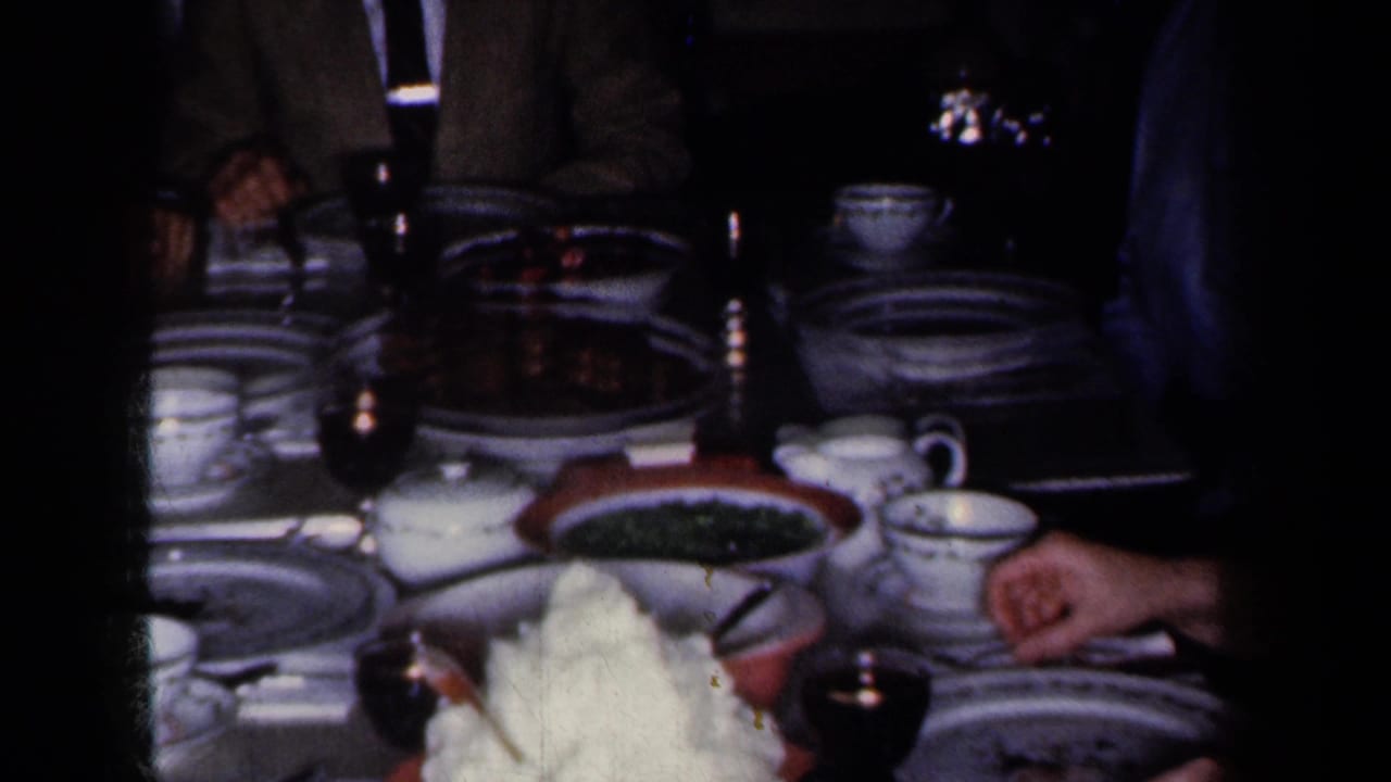 1959 New York home-movie frame of a holiday meal table with china, cups, wine glasses, serving dishes, and adults partly visible around it.