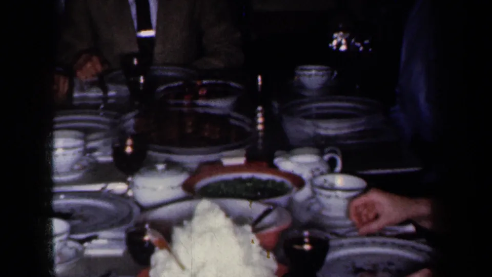 1959 New York home-movie frame of a holiday meal table with china, cups, wine glasses, serving dishes, and adults partly visible around it.
