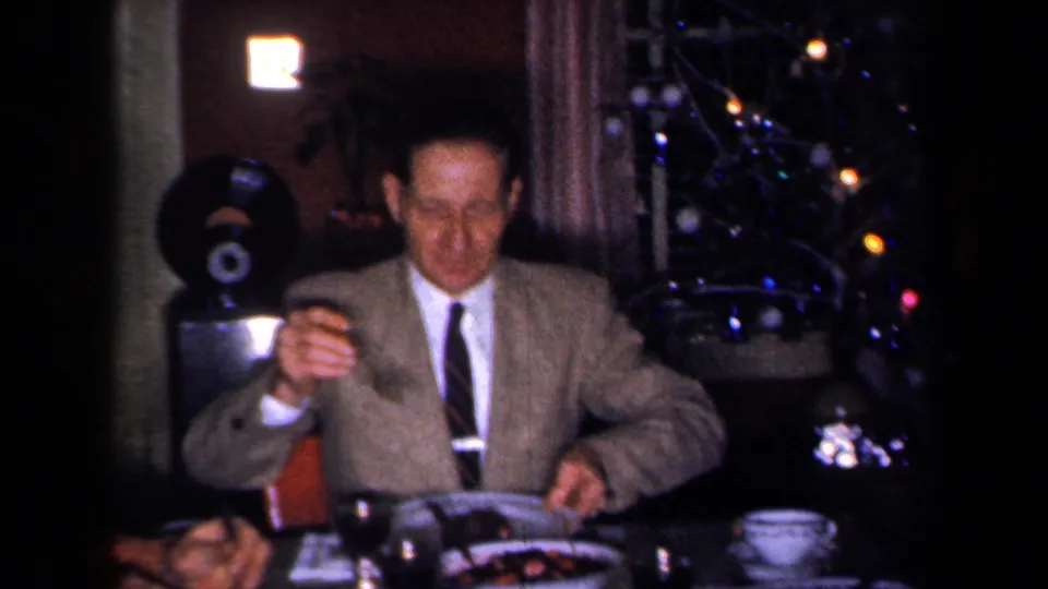 1959 New York home-movie frame showing a suited man seated at a festive dinner table with glassware and a lit Christmas tree in the background.