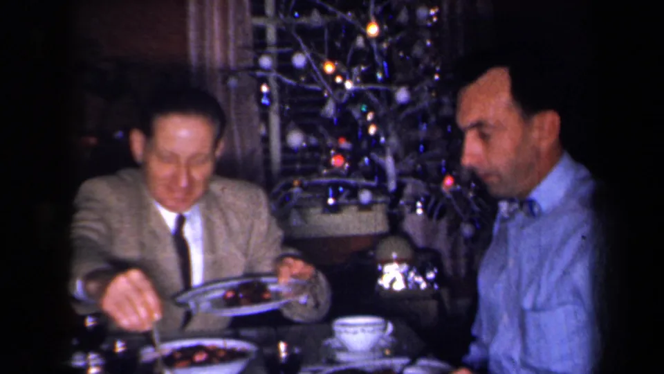 1959 New York home-movie frame showing two adult men at a holiday dinner table, with patterned dishes and a lit Christmas tree behind them.