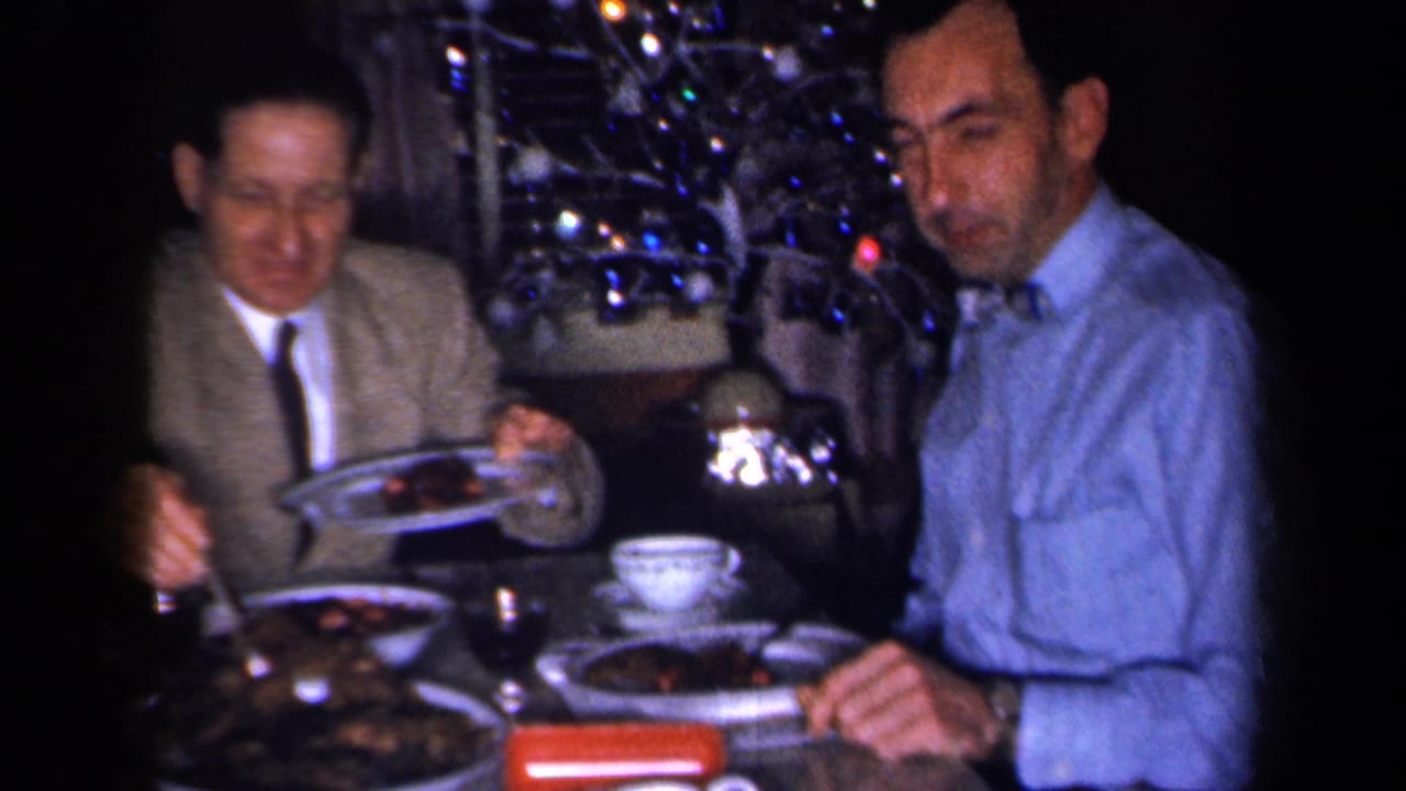 1959 New York home-movie frame showing two men at a holiday dinner table with food, a teacup, and a decorated tree in the background.