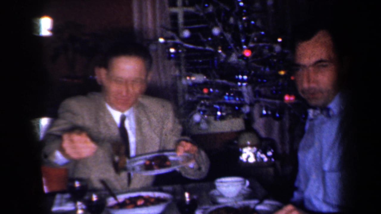 1959 New York home-movie frame of a dinner guest serving food beside another seated man, with a lit Christmas tree in a private interior.