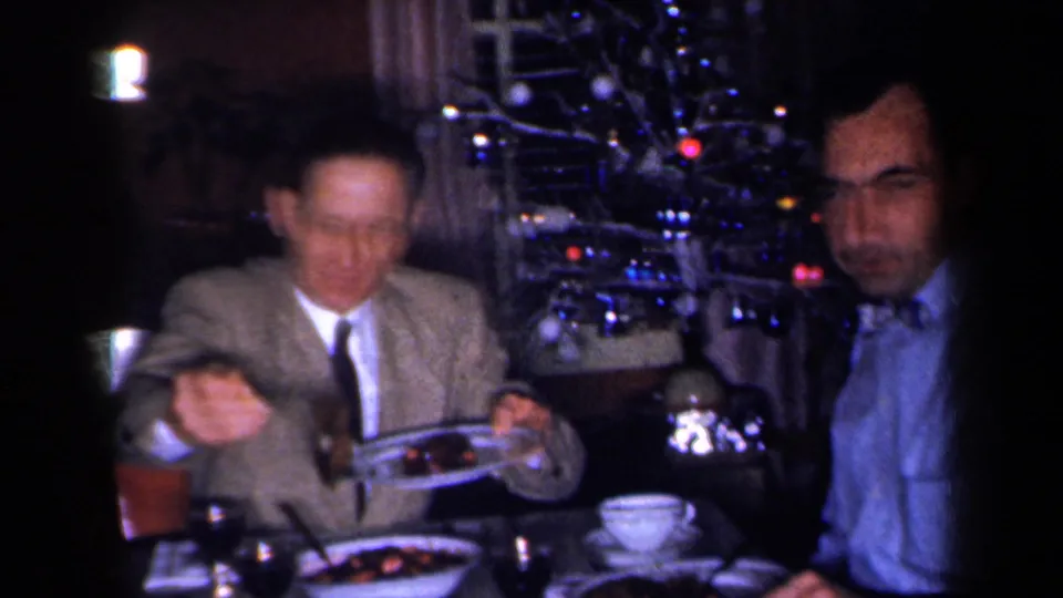 1959 New York home-movie frame of a dinner guest serving food beside another seated man, with a lit Christmas tree in a private interior.