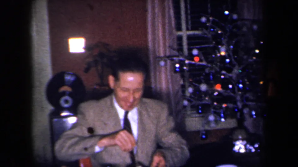 1959 New York home-movie frame showing a suited man at a dinner table with a decorated tree and record player nearby.