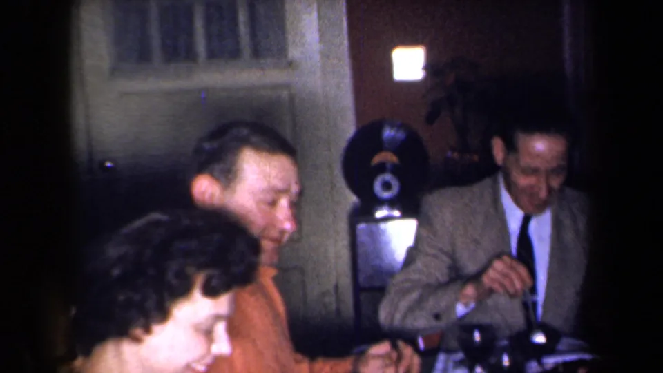 1959 New York home-movie frame showing adults gathered around a holiday dinner table with a record player visible nearby.