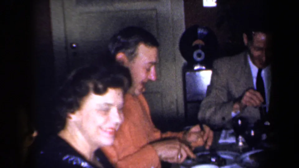 1959 New York 8mm frame showing adults gathered around a dinner table in a dim private interior, with a woman smiling near the camera.