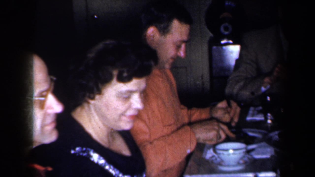 Adults eat at a dinner table, including a woman in a dark dress and a man in an orange sweater.