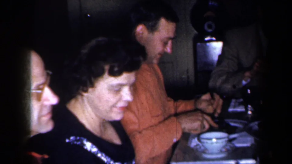 1959 New York home-movie frame of adults eating at a dinner table, with bowls, plates, and a record player-like object in the background.