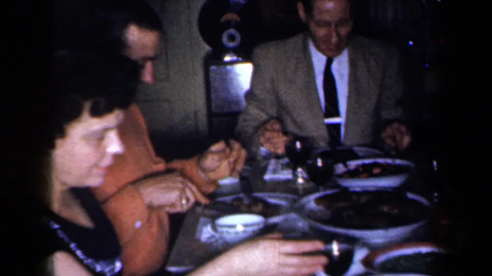 1959 New York 8mm frame showing adults seated around a dinner table in a private room, with serving dishes and tableware in view.