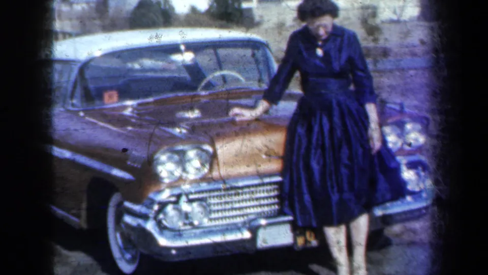 In 1959 New York home-movie footage, a young woman poses beside the front of a gold-and-white car with chrome grille and round headlights.