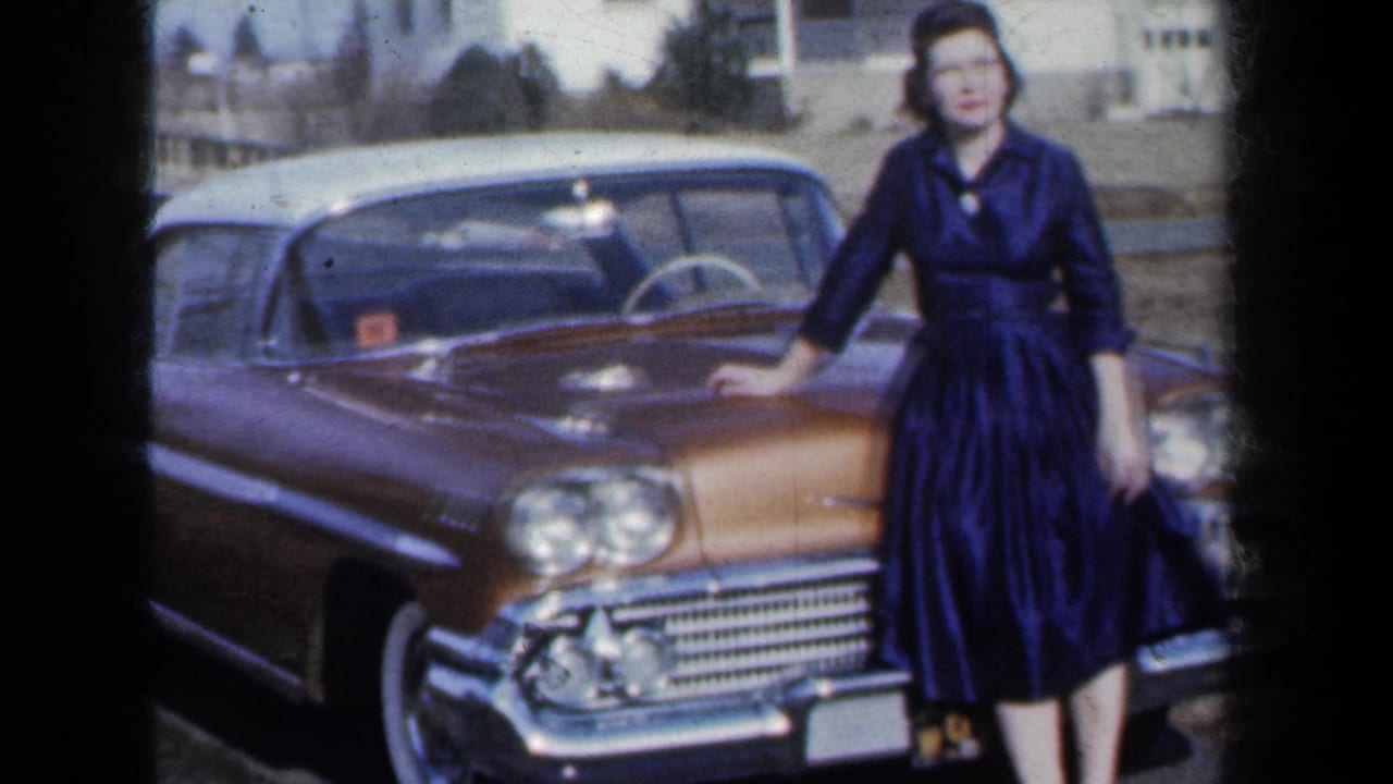 A young woman in a dark dress stands at the front corner of a gold-and-white 1959-era car in a residential New York setting.