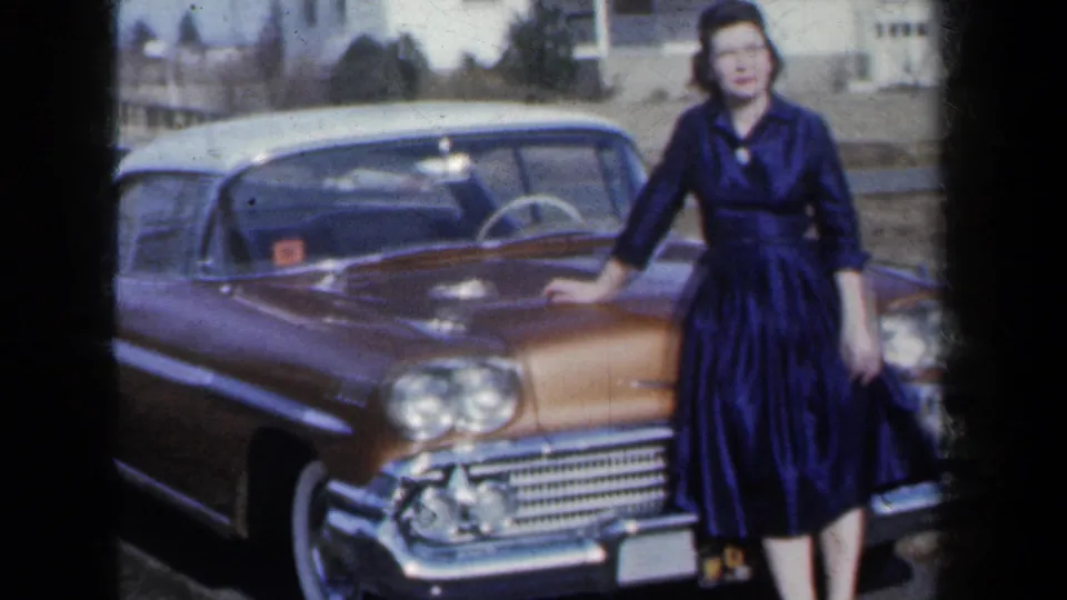 A young woman in a dark dress stands at the front corner of a gold-and-white 1959-era car in a residential New York setting.