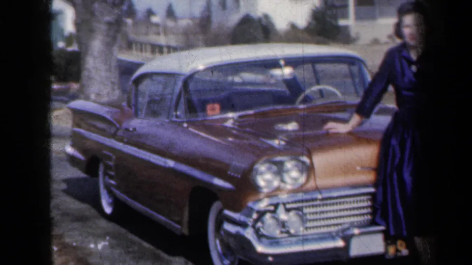 The frame emphasizes the side and front of the gold-and-white car, with the woman partly cropped at the right edge.