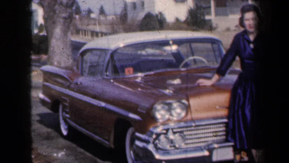 A late-1950s gold-and-white car dominates the frame, with the posing woman partly visible beside the hood in New York home-movie footage.