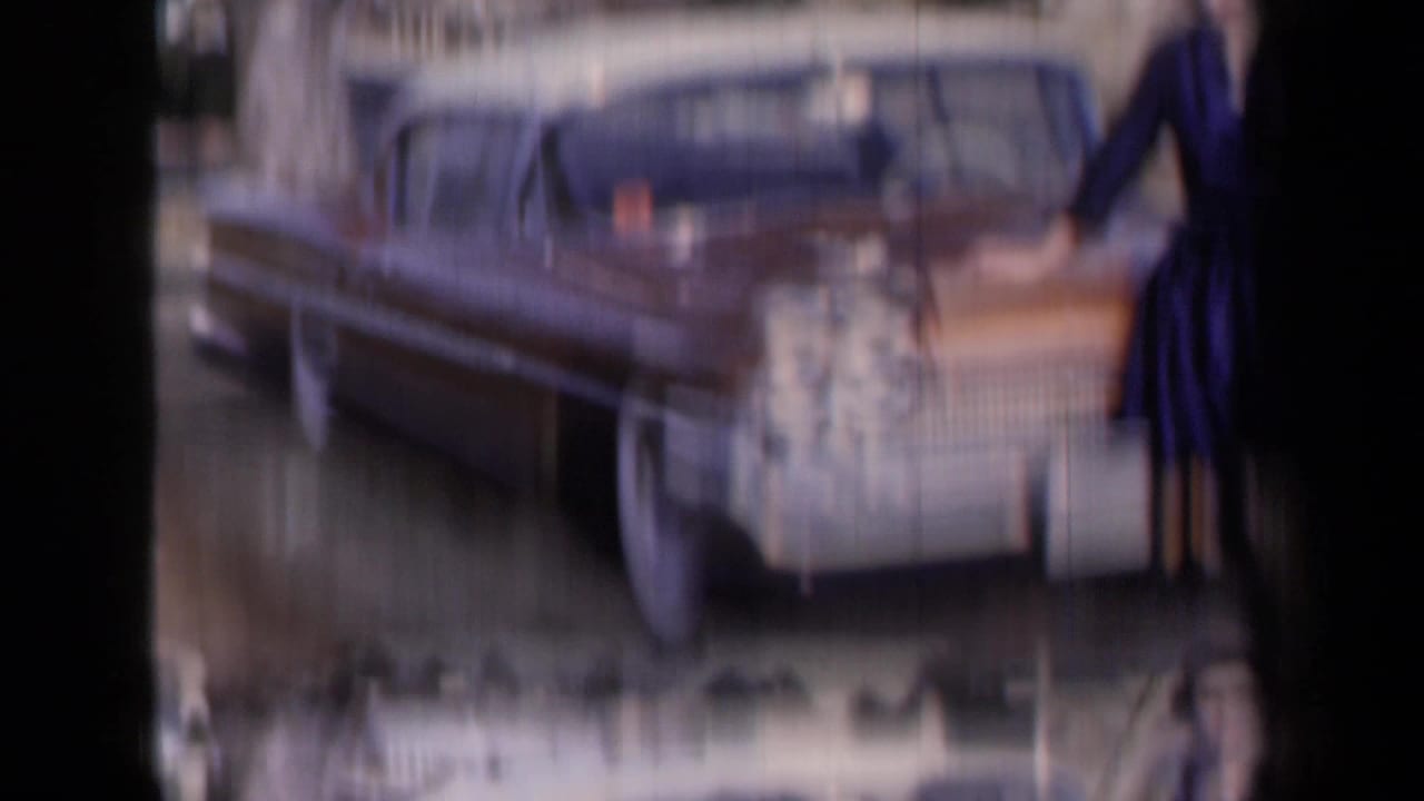 Soft, motion-blurred 8mm frame showing the side of a classic car with part of the woman in a blue dress at the edge.