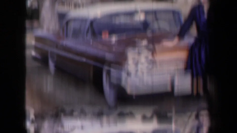 Soft, motion-blurred 8mm frame showing the side of a classic car with part of the woman in a blue dress at the edge.