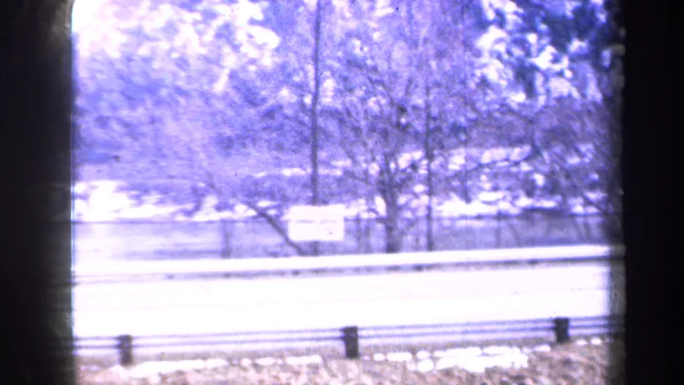 1959 New York travel frame showing a snow-covered roadside, guardrail, trees, and a wooded slope beyond the road.