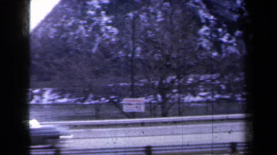 1959 New York home-movie frame of a snowy mountain roadside with a blurred car passing behind a guardrail.