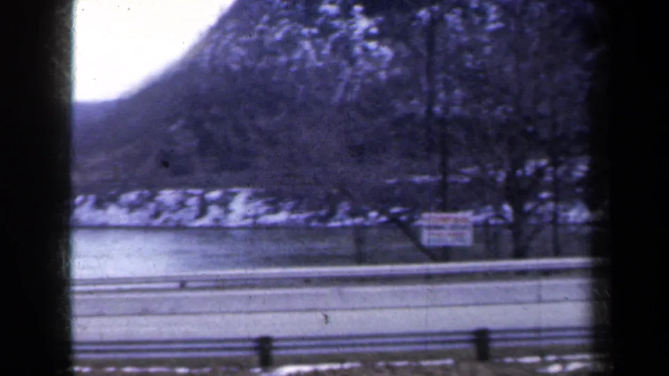 1959 New York 8mm view of a snow-dusted mountain slope, road barrier, water edge, and roadside sign from a moving car.