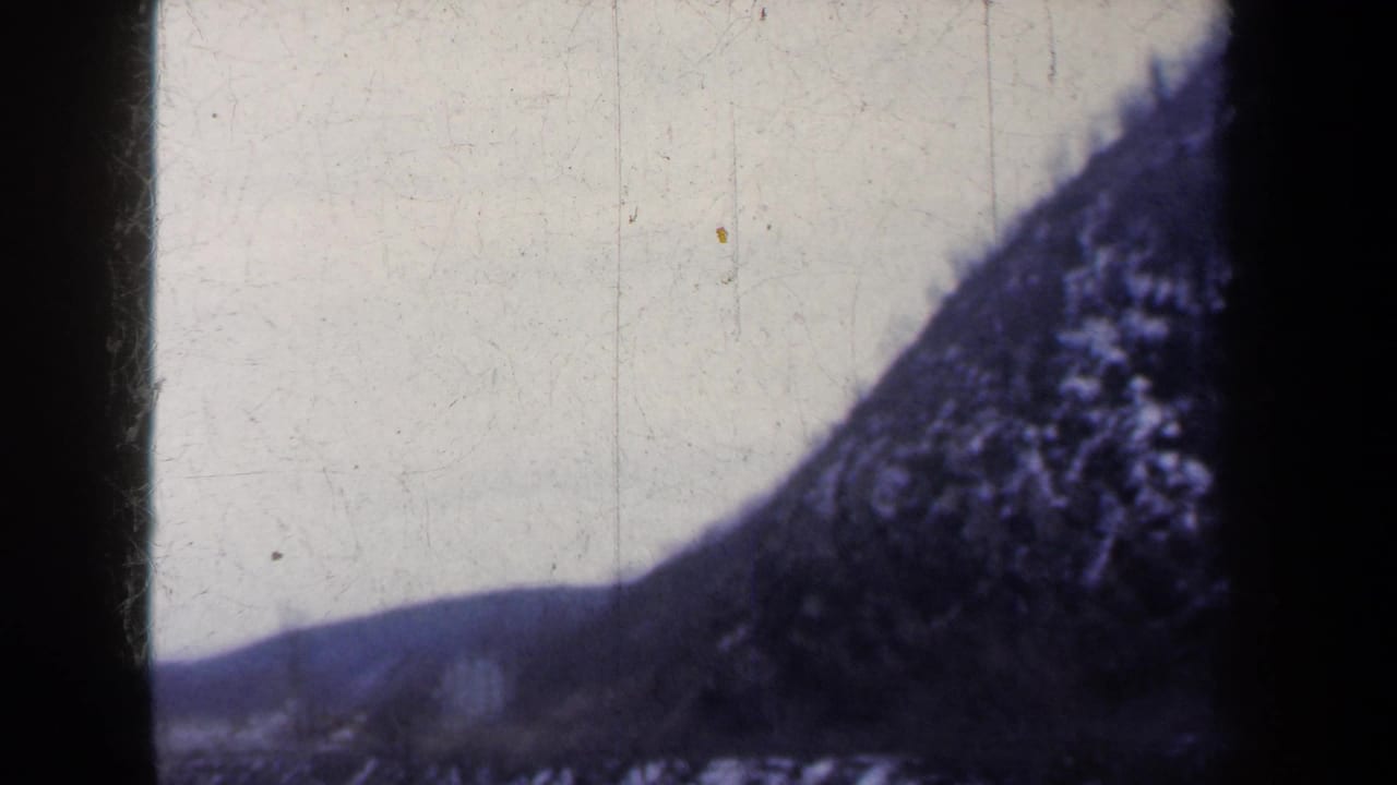 Snow-dusted hillside beside a calm river under a pale sky, framed by dark film borders.