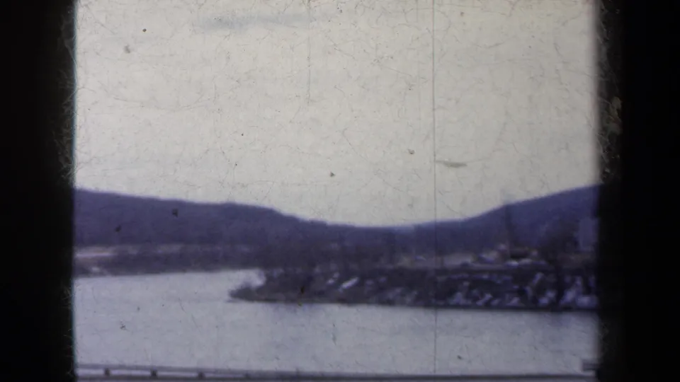 1959 home-movie frame showing a quiet winter riverbank in New York, with snow on the shore and dark hills beyond.